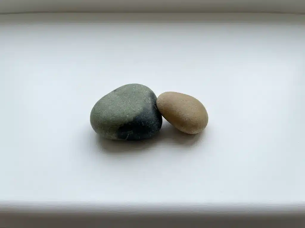Twee rivierkeien naast elkaar op wit oppervlak, één donkerder en groter, één lichter, zachtjes elkaar rakend in minimalistische compositie.