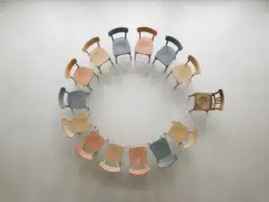 Cirkel van houten stoelen met één losstaande stoel die zich terugbeweegt naar de groep, bovenaanzicht op betonnen vloer.